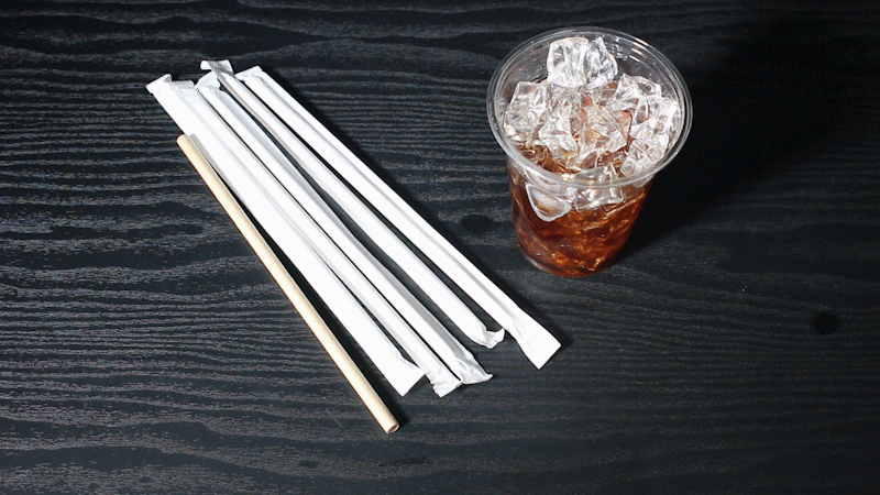 어두운 나무 바닥 배경으로 종이 빨대와 음료가 담긴 플라스틱 투명 컵이 있다. 음료수 컵에 종이 빨대를 꽂는 동작이 반복되는 움직이는 이미지이다.